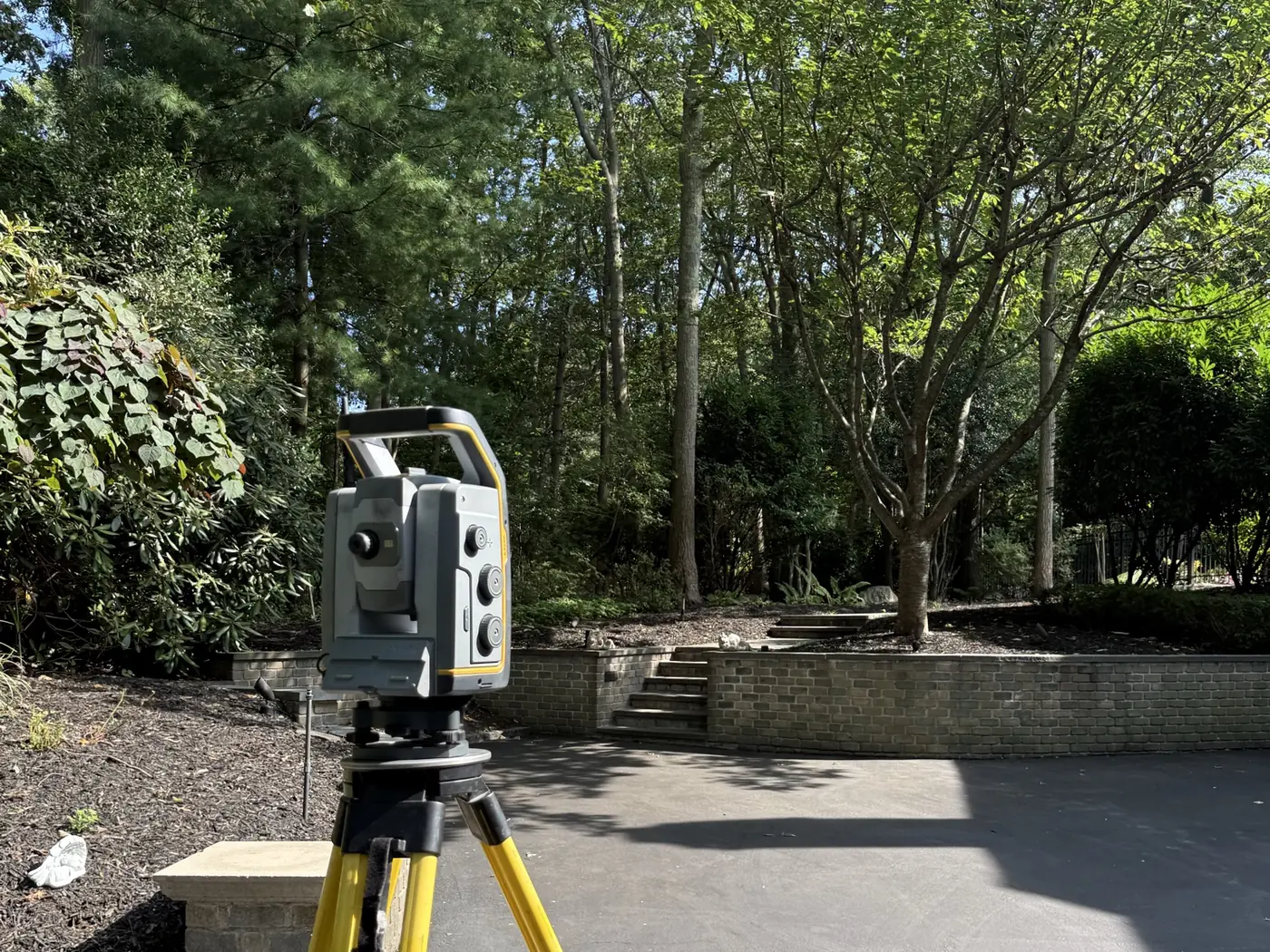 A robotic total station on a yellow tripod occupying a point in a residential driveway, with a brick retaining wall and mature trees behind it