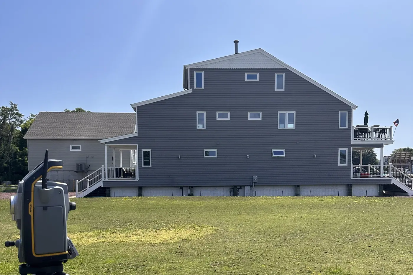 A robotic total station set up on a lawn in front of a large gray waterfront house on a clear day