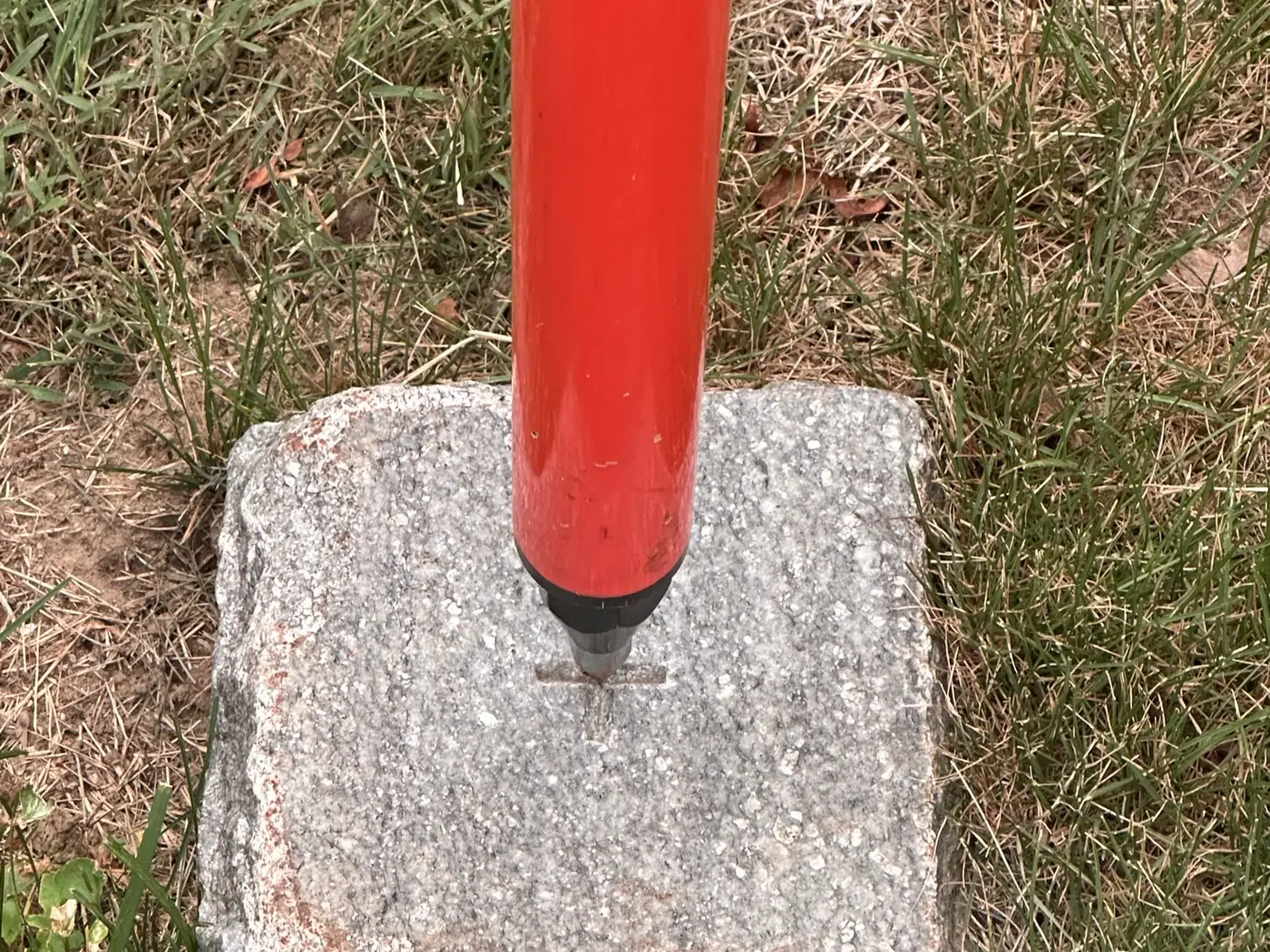 The tip of an orange prism pole held on the drill hole of an existing granite survey monument set in a lawn