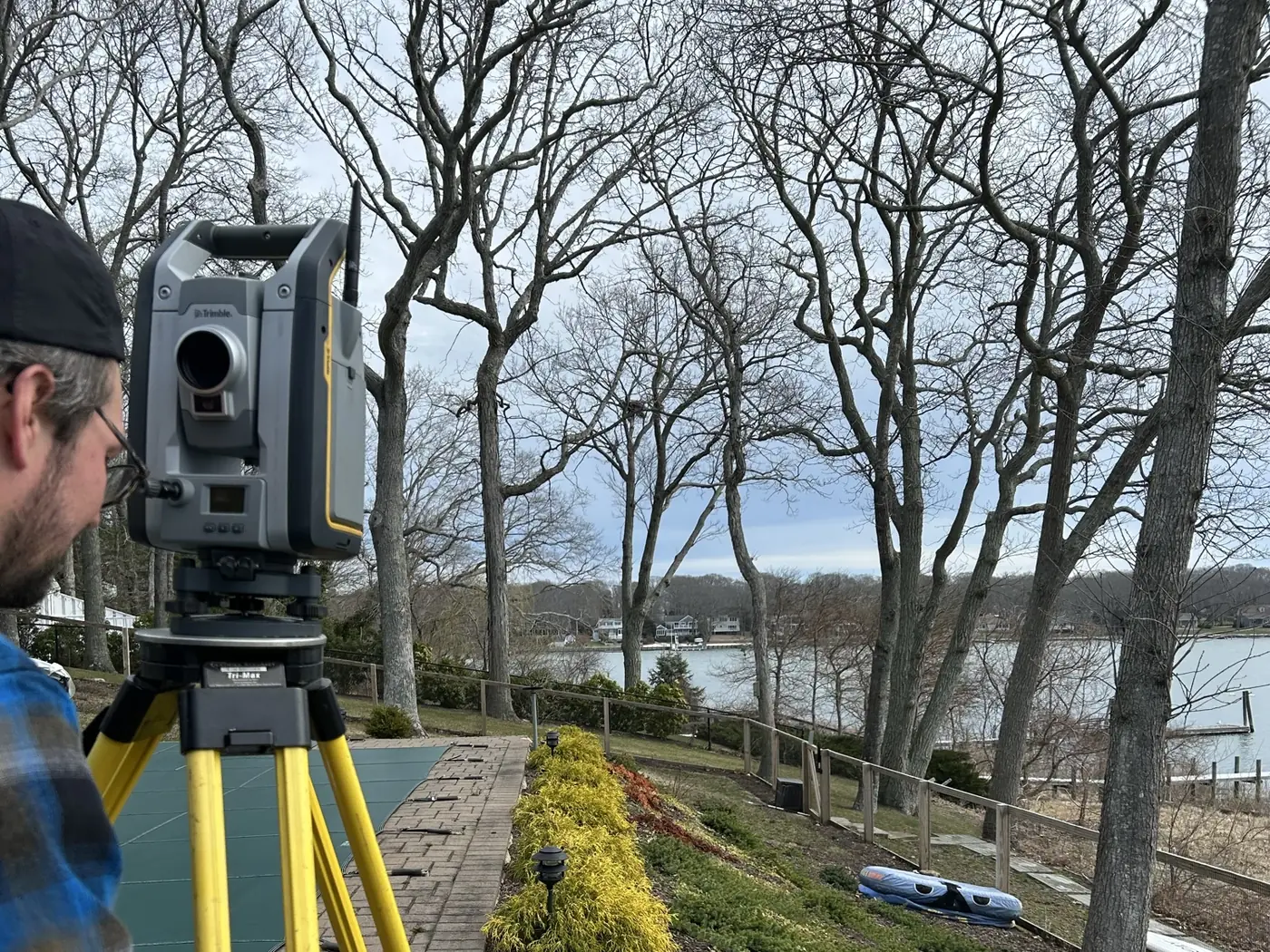 A surveyor sighting through a Trimble total station on a tripod at a waterfront property, with a bay, docks and bulkheads visible through bare trees